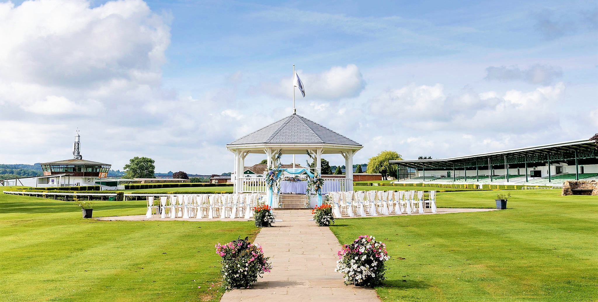 An outdoor wedding ceremony setup at a racecourse featuring a white gazebo decorated with blue and white flowers and ribbons. White chairs are arranged neatly on either side of a stone pathway leading up to the gazebo, surrounded by green lawns and floral planters under a bright blue sky.